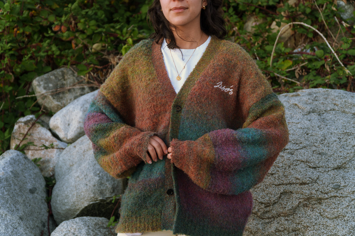 A person outdoors wearing a fuzzy, oversized cardigan with a mottled color palette of green, brown, orange, and purple. The cardigan features a white embroidered script logo on the left chest and is worn over a white t-shirt, set against a background of rocks and green foliage.