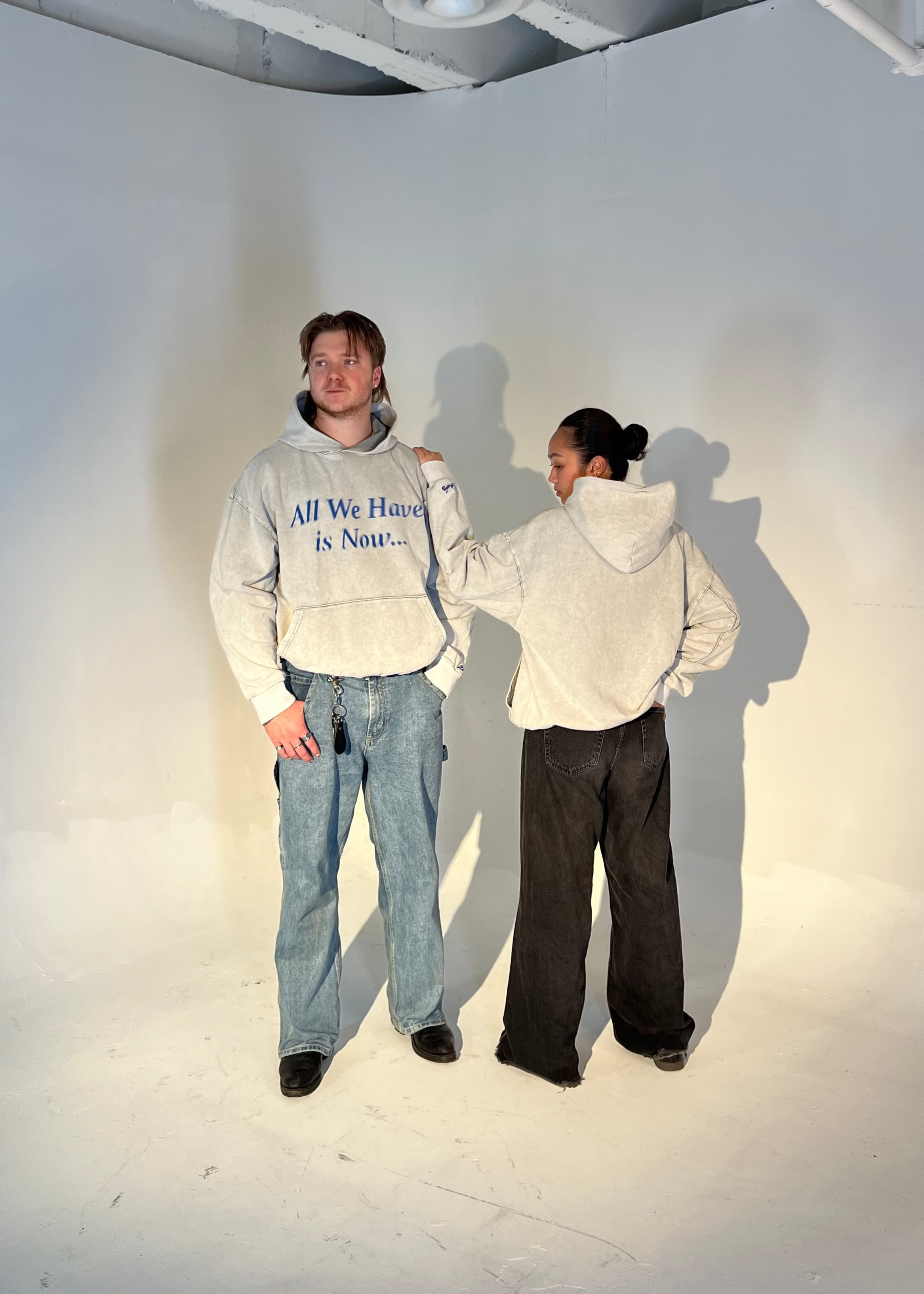 Two people standing in a studio setting, one with a hoodie featuring the text 'All We Have is Now' on it, 

#zebos #ootd #aesthetic #clothingbrand #luxurygarment #streetwear #fashionstreetwear #fashiondesigner #pinterest #outfitinspo #falloutfits #autumnfits #cleanfits #oversized #baggyfits #newcollection #newdrop #bts #ceo #fashionbrand #brandowner #entrepreneur #fashionbusiness #motivation#fw25 #necktag #smalldetails
